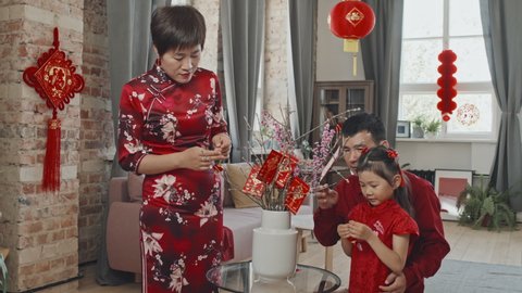 Medium shot with slowmo of Chinese family in red traditional clothing putting Lunar New Year ornaments and postcards on branches standing in vase in their living room decorated with paper lanterns