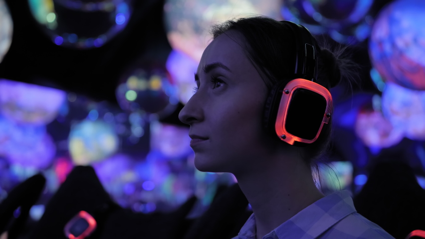 Close up: woman wearing wireless black headphones and looking around in dark room of interactive museum or exhibition with colorful illumination. Futuristic, immersive, entertainment concept