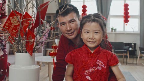 Portrait shot of happy Asian father and cute 5-year-old girl in traditional dress putting ornaments on branches in vase while decorating for Chinese New Year, then smiling for camera