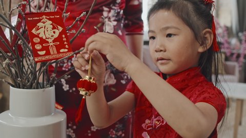 Close up slowmo of cute Chinese little girl in red traditional dress putting Lunar New Year ornaments on branches in vase while her unrecognizable mother in qipao helping her