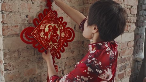 Portrait with close up of happy Chines woman in qipao hanging Lunar New Year ornament on wall, then smiling for camera