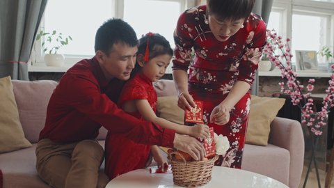 Medium shot of Asian mother in traditional dress and father in red shirt helping cute 5-year-old girl preparing gift basket for Chinese New Year celebration