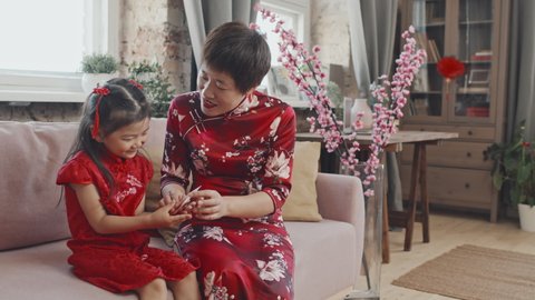 Medium shot with slowmo of happy Asian mother in traditional dress giving red postcard to cute 5-year-old girl sitting on couch in living room decorated for Chinese New Year