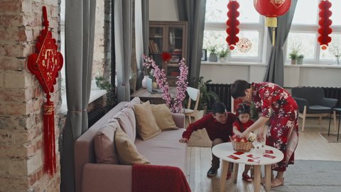 Medium shot of Chinese family with little daughter wearing traditional clothing and arranging gift basket for Lunar New Year in living room decorated with paper lanterns and ornaments