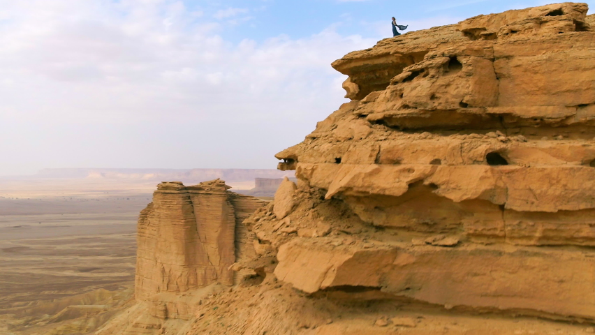 Aerial Panning Over A Woman Standing On The Edge Of A Soaring Cliff And Holding Her Scarf In The Breeze - Arabian Desert, Saudi Arabia