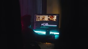 Teenager using computer to edit footage while sitting at desk in obscure room at night at home