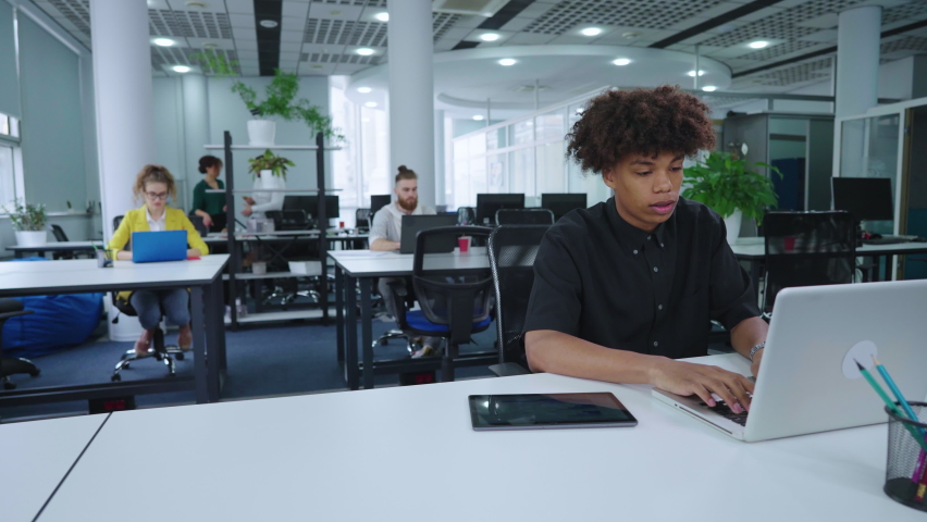 African American manager working on laptop in office, swiping reports on statistics on tablet, diverse coworkers on background. Arc shot young analyst doing research. Concept of business