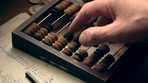 Male hand counting money using old wooden abacus with old paper with mathematical calculations and metal coins on the table close up