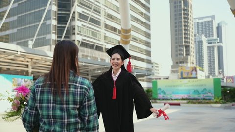 Happy beautiful young woman in graduation gowns with holding a diploma and hugging mother by love and proud in city. Asian mom embraces daughter with joy on graduation day and successful. Slow motion.