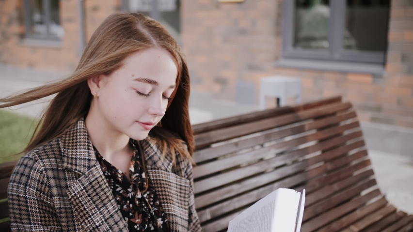 A young girl with blonde hair reads a book on a bench