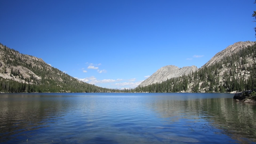 A beautiful, blue sky summer day at Toxaway Lake, a large alpine lake in the Sawtooth Mountains near Stanley, Idaho. The water surface is tranquil, a few clouds are seen in the distance.