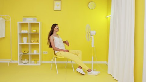 Young beautiful woman, model in yellow trousers, white top and cat eye glasses, is posing on yellow chair in front of electric fan, blowing her long hair, she is trying to cool down in hot office.