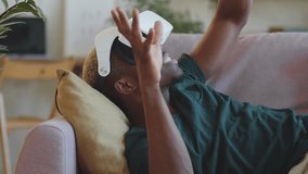 Delightful young African American man lying on sofa, smiling and touching something in air while experiencing augmented reality with VR glasses at home