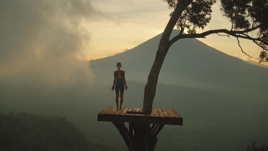 Slender woman on wooden tree platform raising arms into greeting pose, mount Agung Royalty-Free Stock Footage #1081828193