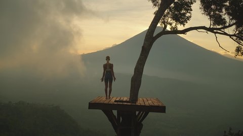 Slender woman on wooden tree platform raising arms into greeting pose, mount Agung