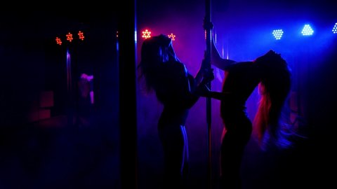 Two slender young women dance near a pole in high-heeled shoes. Silhouette of dancers in a nightclub.