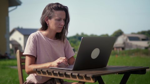 Female freelancer in casual clothes sit at the desk on the backyard of her house and works remotely with laptop computer. Distance work in the garden at sunny day