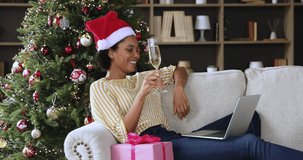 Female sit on sofa at home join virtual New Year party in video chat on laptop wave hand raise champagne glass celebrate Xmas. Young Black lady in Santa hat meet Christmas with mates online