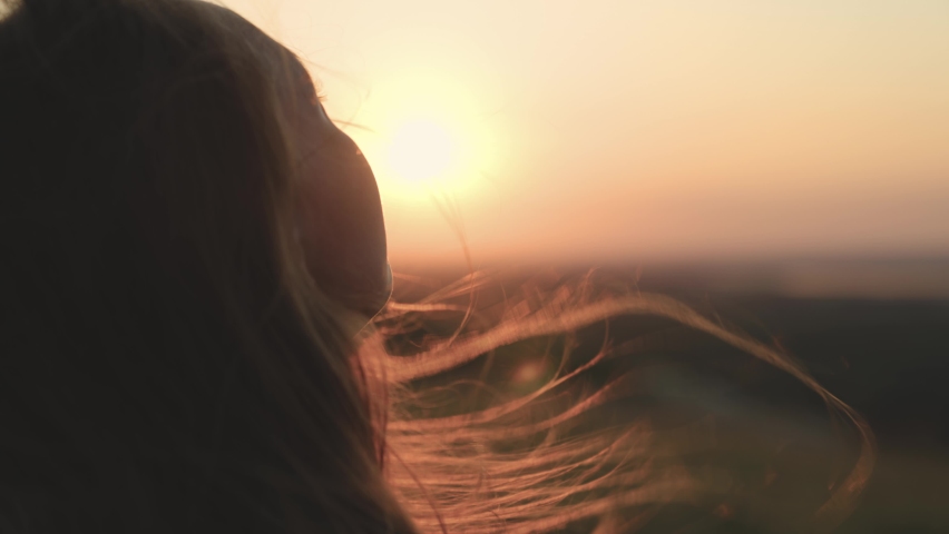 Girl prays looking at sunset, long hair is flying away in glare of sunlight rays in strong wind, looking at dawn, lonely hike of brave girl, looking into sky with her eyes, believing good
