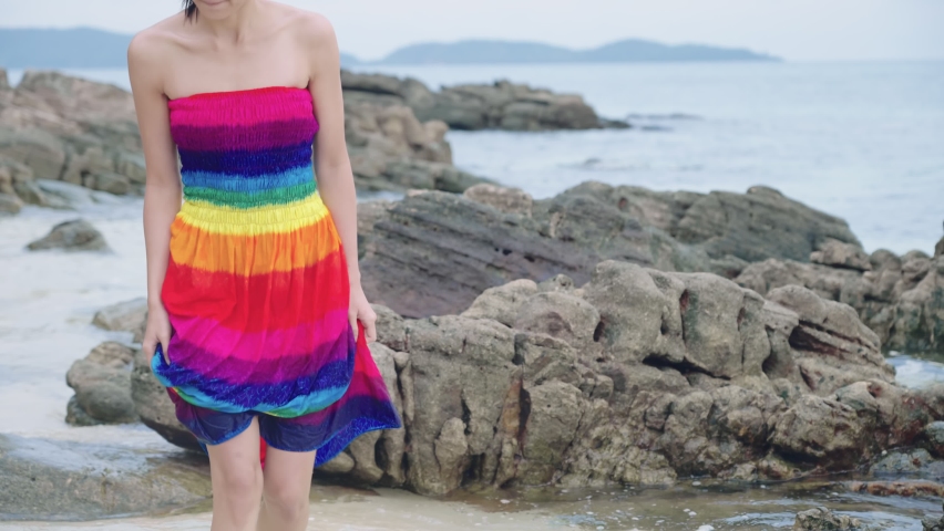 A female tourist body getting cold  in a wet rainbow dress while walking against ocean breeze on a white rocky beach, tropical island exploring activity, relaxing exercise, touching natural resources