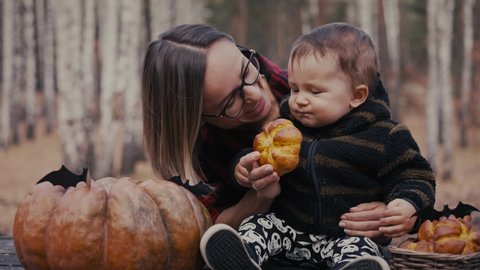 Young woman and her baby son in autumn park, boy playing with helloween pumpkin and eating pumpkin bun