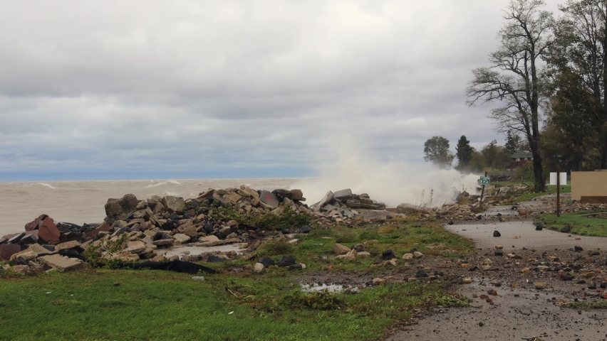 Waves crashing into rocks on edge of Lake Michigan.  Cloudy sky and with wisps of blue.  Sidewalk covered with rocky debris.  Directional sign at angle from wind.  Recreational trail and fence.