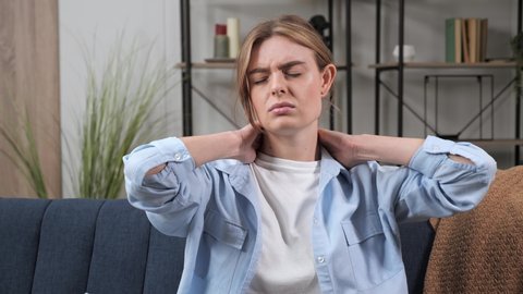 Young caucasian woman in a white T-shirt and a blue shirt sitting on the couch in the living room holding her low back, feeling severe pain in it. Cervical osteochondrosis, tired overworked female.