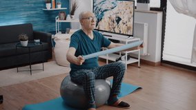 Senior man using resistance band to do physical exercise with arms while sitting on fitness toning ball. Aged person training with flexible elastic belt and workout equipment. Adult exercising