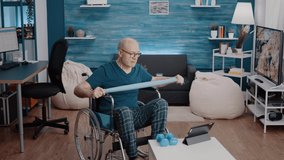 Old person with disability pulling resistance band to stretch while following workout video online on tablet. Senior man sitting in wheelchair and watching training lesson on device.