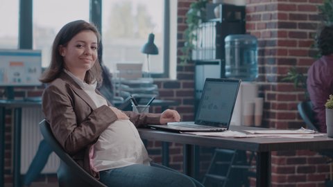 Portrait of pregnant woman working with charts on laptop. Employee expecting child and planning business project with documents and computer at startup office. Adult doing paperwork