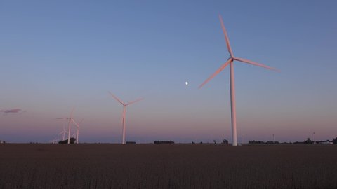 AMERICA - CIRCA 2020s - The moon rises behind windmills turning on the flat farmlands of central Indiana and Illinois generating clean energy.