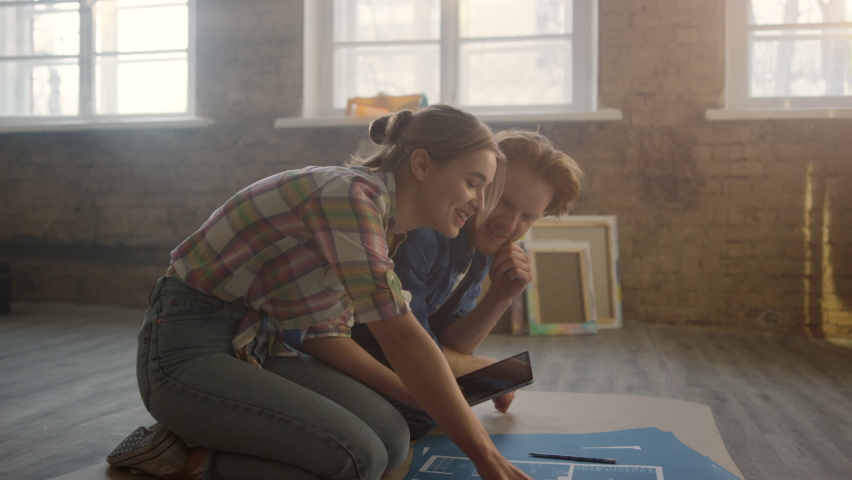 Smiling people looking to blueprints of new house on floor. Cheerful family working with tablet on design project in light room. Happy couple planning home repair together indoors. Royalty-Free Stock Footage #1081845656