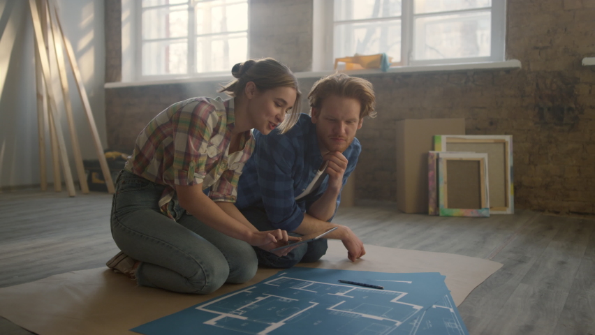 Smiling people looking to blueprints of new house on floor. Cheerful family working with tablet on design project in light room. Happy couple planning home repair together indoors.