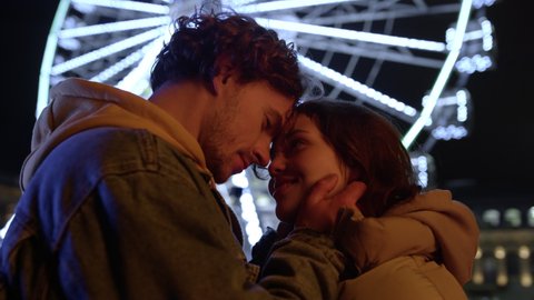 Smiling couple looking eyes to eyes on illuminated ferris wheel background. Affectionate man touching woman face outdoor. Lovely pair felling happy together on urban street. Romantic love concept.