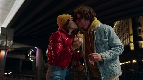 Happy couple kissing with snacks in hands under bridge. Hungry man biting off woman burger on city street at night. Playful girlfriend and boyfriend having fun outdoor during romantic date.