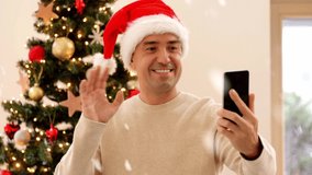 technology and witer holidays concept - happy man in santa hat with smartphone having video call and showing christmas tree at home over snow