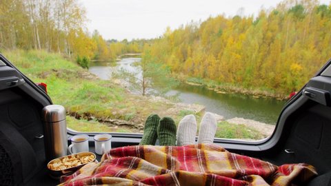 travel, tourism and camping concept - view to river from car trunk with feet in warm socks under blanket, two cups of tea, thermos and cookies