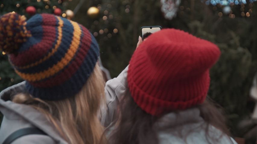 Two happy girls use smartphone at Christmas  decorated town square with fair lights in background and wave hands to smiling male friend on screen. Celebrating Christmas New Year online. Together joy Royalty-Free Stock Footage #1081856468