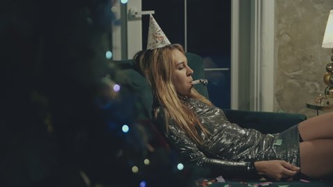 Side view of upset blonde girl lying in chic dress on couch or sofa and feeling lonely. Sad woman alone in party or birthday cap blowing whistle and celebrating birthday, New Year, Christmas or Xmas