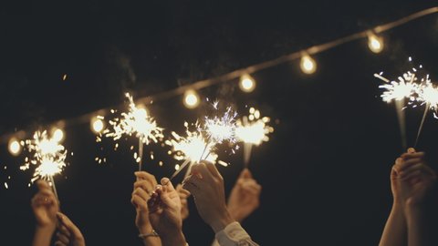 Close-up of hands or palms holding and waving burning Christmas sparklers in front of black or dark background. Sparkling lights at birthday party, wedding, New Year, Christmas Eve, Xmas. - Βίντεο στοκ