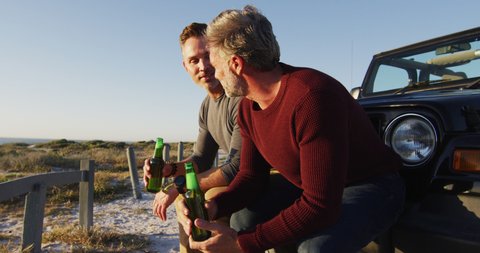 Happy caucasian gay male couple sitting by car drinking bottles of beer on sunny day at the beach. summer road trip and holiday in nature. Stock Video