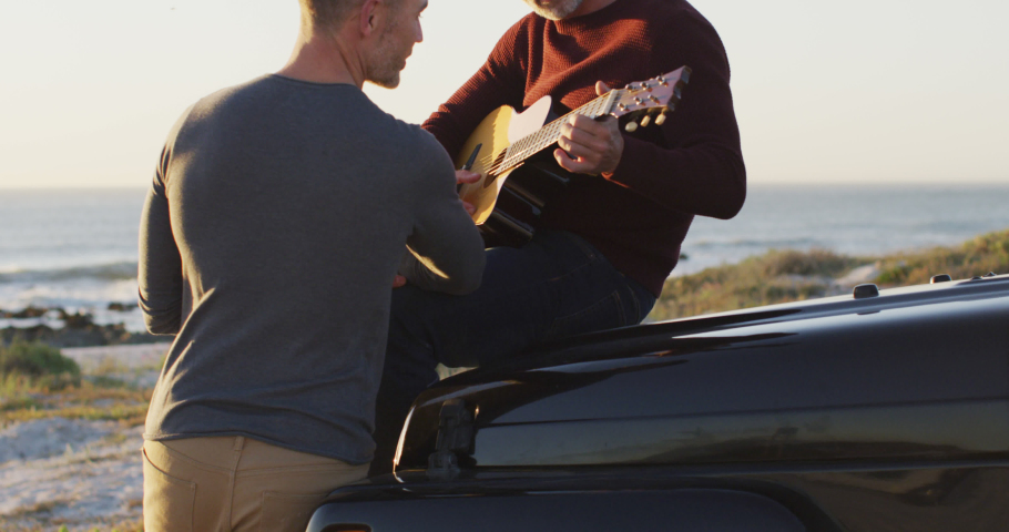 Happy caucasian gay male couple sitting on car playing guitar and talking at the beach. summer road trip and holiday in nature.