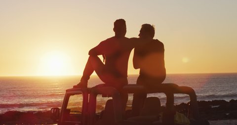 Happy caucasian gay male couple sitting on car roof embracing at sunset on the beach. summer road trip and holiday in nature. 스톡 비디오