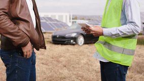 Details closeup taking video of ecological engineer and ecological worker together end the discussion at solar panels farm and continue to walking to the electric car beside of photovoltaic batteries