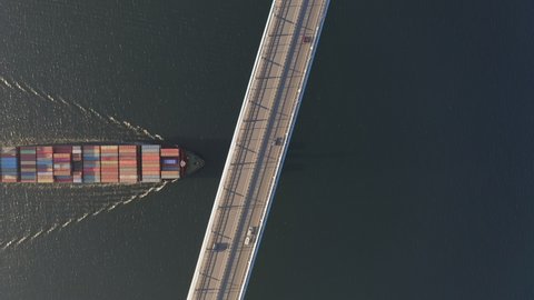The container ship passes under a large bridge over which cars are moving. The vessel is transporting containers. Waves radiate from the bow of the ship. View from the drone vertically down