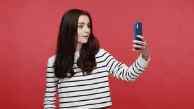 Woman holding smartphone in hand and posing to camera, making selfie photo for social networks, wearing casual style long sleeve shirt. Indoor studio shot isolated on red background.