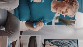 Vertical video: Senior adult doing arm stretch with dumbbells on fitness toning ball. Aged woman using weight lifting while following video of online training lesson to practice exercise at home.