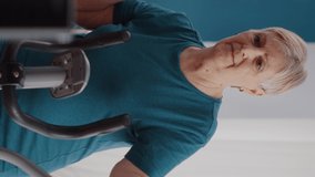 Vertical video: Close up of retired woman riding stationary bicycle at home. Senior person using cardio machine to cycle and do physical exercise on static bike. Pensioner cycling and doing fitness