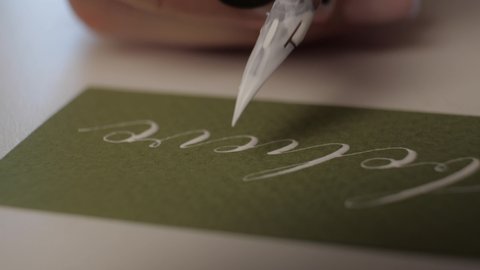 close-up calligraphy. woman applies white paint with nib of a pen on green paper. the girl slowly writes the word believe in beautiful handwriting. penmanship