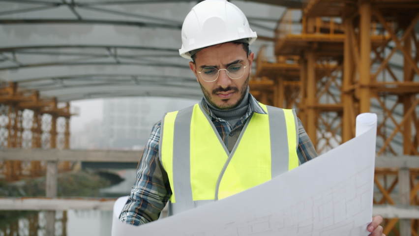 Serious Arab guy engineer is checking construction plan and looking around outdoors in industrial zone. Building specialists and workplace concept. | Shutterstock HD Video #1081888178